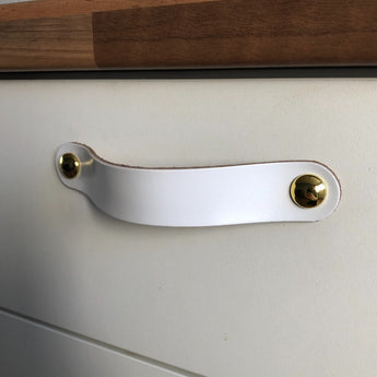 Een close-up van een Tomasso White lade van yessential.nl, met wit lederen handgrepen die worden vastgezet door twee gouden ronde sluitingen, onder een houten aanrechtblad.