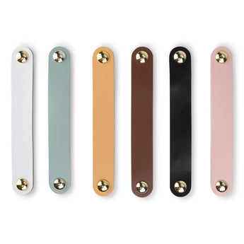 Zes platte, rechthoekige Tomasso White lederen handgrepen van yessential.nl, in wit, lichtblauw, tan, bruin, zwart en roze met afgeronde uiteinden en gouden sluitingen aan beide uiteinden, verticaal naast elkaar geplaatst op een witte achtergrond.