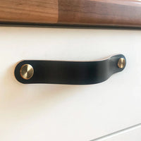 Een close-up van een lade met de Luciano Black Deluxe greep van yessential.nl: een luxe zwart lederen greep met twee messingkleurige sluitingen tegen een wit oppervlak en een houten bovenrand.