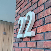Het stijlvolle huisnummer Enrico beton 20cm van yessential.nl is duurzaam en gemonteerd op een rode bakstenen muur naast een deur met houten panelen en een vierkante lamp.