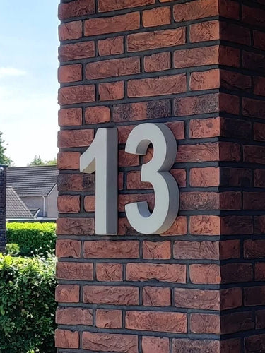 De Enrico beton 30cm van yessential.nl is een vet wit nummer 13 in 3D huisnummer stijl, gemonteerd op een rood en donkerbruine bakstenen zuil buiten een gebouw, met groen en naburige huizen op de achtergrond.