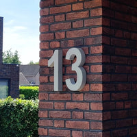 De Enrico beton 30cm van yessential.nl is een vet wit nummer 13 in 3D huisnummer stijl, gemonteerd op een rood en donkerbruine bakstenen zuil buiten een gebouw, met groen en naburige huizen op de achtergrond.