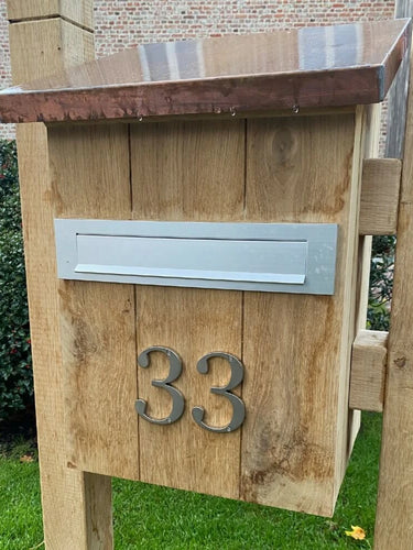 Een houten brievenbus met een metalen brievenbus en yessential.nl Matteo Nickel 10cm huisnummer 33 in geborsteld ontwerp op de voorkant, staand op een grasveld met struiken en een bakstenen gebouw op de achtergrond.