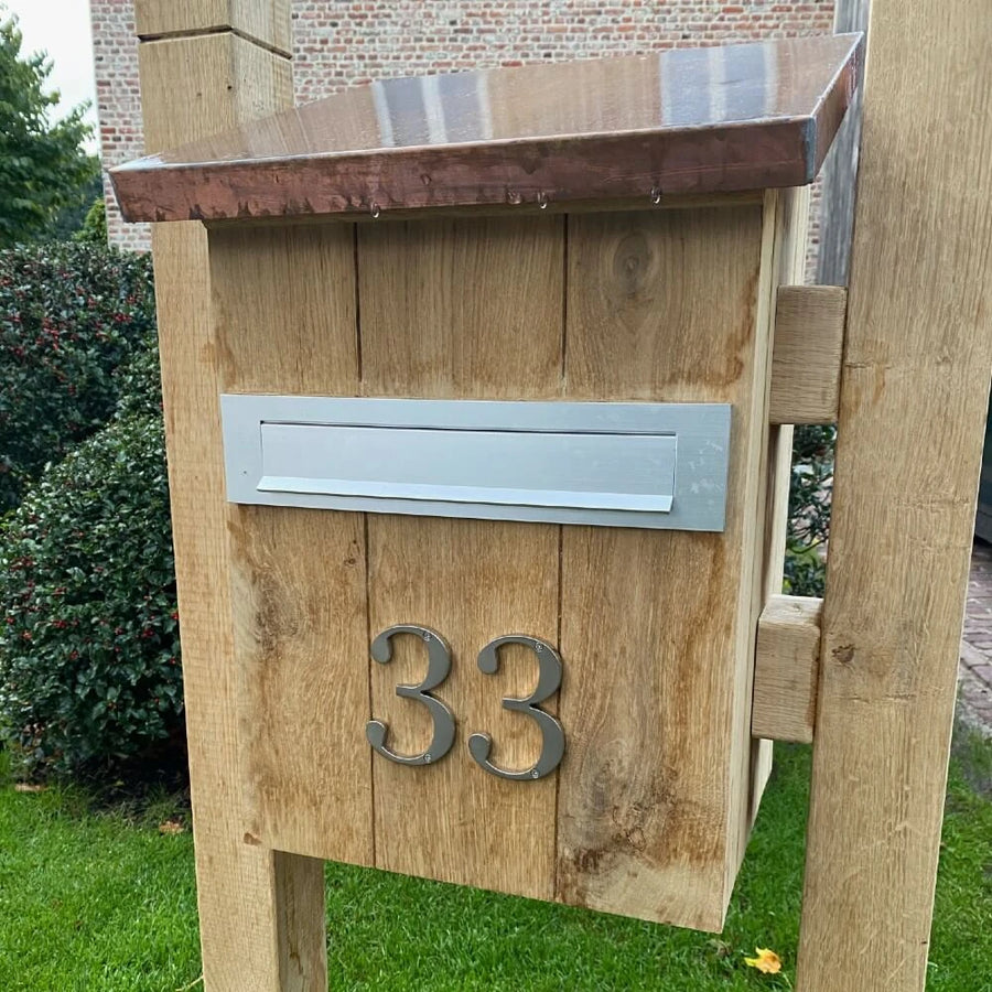 Een houten brievenbus met een metalen brievenbus en yessential.nl Matteo Nickel 10cm huisnummer 33 in geborsteld ontwerp op de voorkant, staand op een grasveld met struiken en een bakstenen gebouw op de achtergrond.