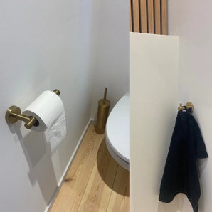 Een kleine moderne badkamer met een licht houten vloer, een wit toilet, gouden toiletpapierhouder en borstel, en de Justo Gold Hook van yessential.nl die een zwarte handdoek vasthoudt voor een elegante touch.