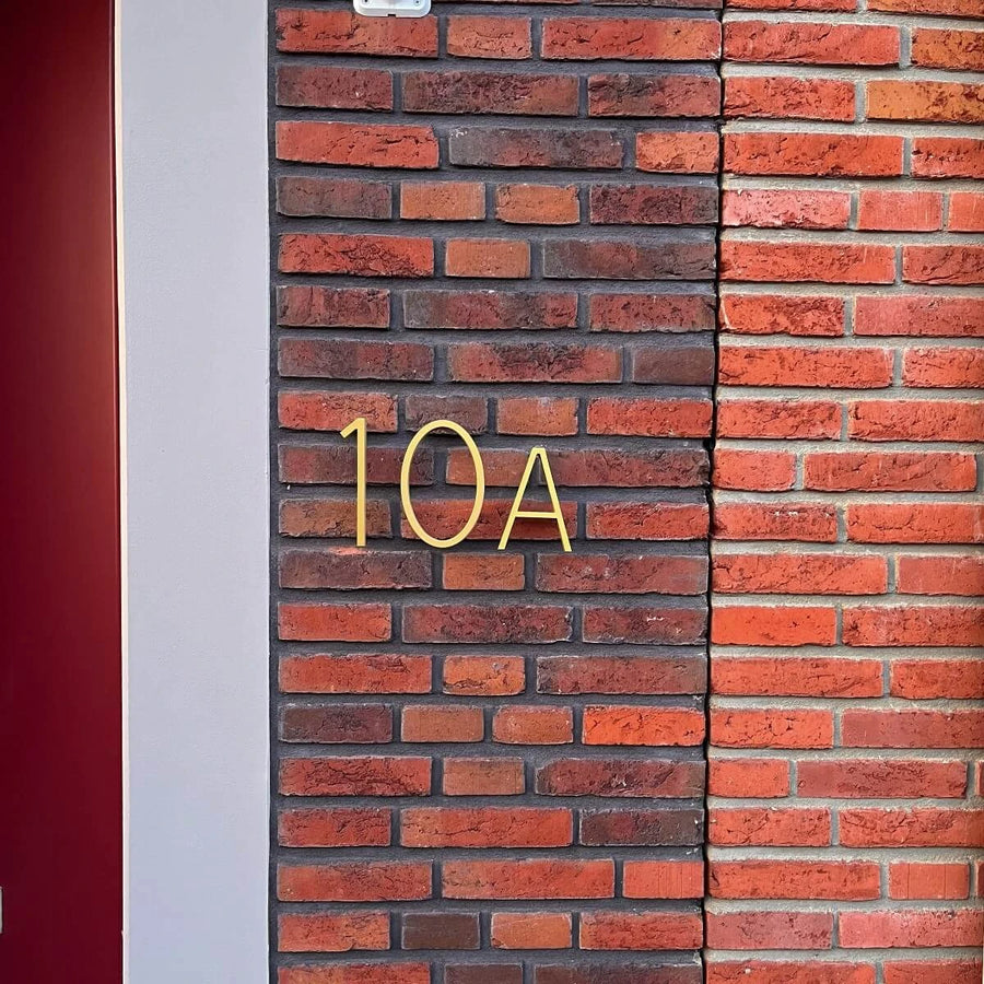 De Amato Gold 15cm nummer en letter van yessential.nl zijn vormgegeven als moderne huisnummers en gemonteerd op een bruin en rode bakstenen muur naast een lichtgrijze verticale strip en een rood deurkozijndeel aan de linkerkant.