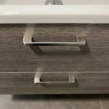 Een close-up van twee Ramiro Chrome Deluxe handgrepen van yessential.nl op een lade van een badkamermeubel met houtstructuur, met een witte wasbakrand erboven en strakke grijze tegels op de vloer.