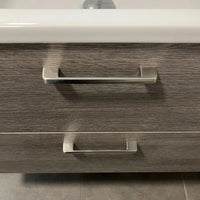 Een close-up van twee Ramiro Chrome Deluxe handgrepen van yessential.nl op een lade van een badkamermeubel met houtstructuur, met een witte wasbakrand erboven en strakke grijze tegels op de vloer.