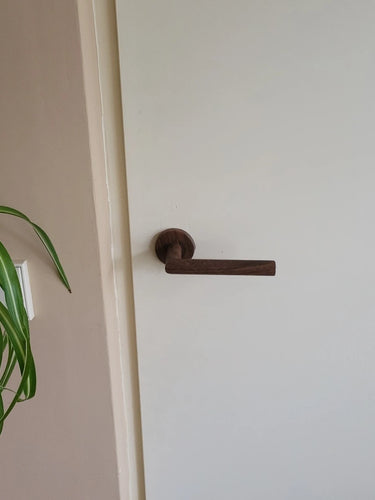 Een witte deur met een donkere, moderne Lucio Walnut deurklink binnendeur van yessential.nl. Links zijn een groene bladplant en een lichtschakelaar op een beige muur zichtbaar.