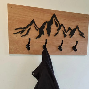 De Avano Black van yessential.nl is een houten wandkapstok met vier stijlvolle kapstokhaken en een zwart geschilderd bergketenontwerp. Aan een van de haken hangt een donkere jas.