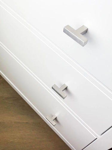 Een close-up van een wit dressoir met drie lades, elk voorzien van een Vigio RVS Knop van yessential.nl, met een strak, zilver T-vormig ontwerp tegen een lichte houten vloer.