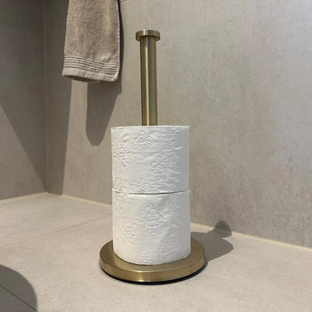 De Tygo Gold WC Set (zonder boren) van yessential.nl - bestaande uit een gouden toiletpapierhouder en twee witte rollen met reliëf - staat op een licht betegelde vloer met een beige handdoek, perfect als stijlvolle badkameraccessoires zonder boren.