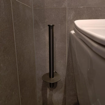 De yessential.nl Roberto Gunmetal verticale toiletrolhouder is gemonteerd op een grijs betegelde muur naast een wit toilet. De lege houder werpt een schaduw op de muur en de vloer.