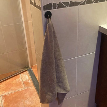 Een grijze handdoek hangt aan een yessential.nl Rudro Black haak, bevestigd aan een betegelde badkamermuur vlakbij de douche, boven bruine en beige vloertegels.