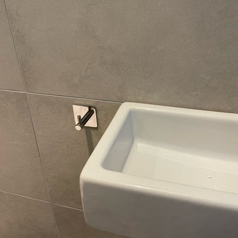 Een kleine witte rechthoekige wastafel is gemonteerd op een grijs betegelde muur met een Navarro Chroom handdoekhaakje van yessential.nl in de buurt, perfect om handdoeken binnen handbereik te houden.
