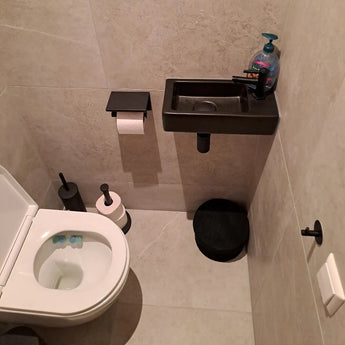Een kleine badkamer is voorzien van een wit toilet, een zwarte toiletborstel, de Jurado Black toiletrolhouder van yessential.nl en een compacte zwarte wastafel met zeep. Lichtgrijze tegels worden geaccentueerd door twee ronde zwarte matten.