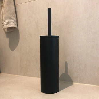 De Bruno Black vrijstaande toiletborstel van yessential.nl met cilindervormige houder staat op een lichte badkamervloer tegen een betegelde muur, met een opgevouwen handdoek in de buurt.