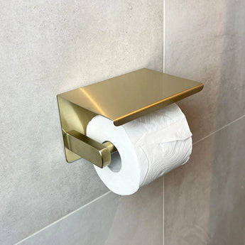 Een rol toiletpapier hangt aan een gouden houder uit de Armando Gold Set van yessential.nl, gemonteerd op een lichtgrijs betegelde badkamermuur.