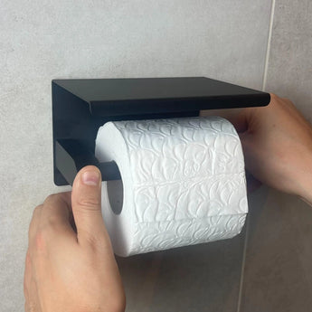 Iemand plaatst een rol wit toiletpapier met reliëf op de Tygo Black WC Set (no-drill) van yessential.nl, die zonder boren is gemonteerd op een lichtgrijze betegelde muur.
