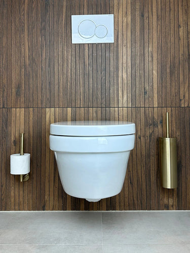 Een moderne badkamer toont een aan de muur gemonteerd wit toilet tegen houten lambrisering, met rechts de Fabiano Gold WC-set van yessential.nl, inclusief een gouden toiletborstelhouder. De vloer en zijwanden zijn lichtgrijs betegeld.