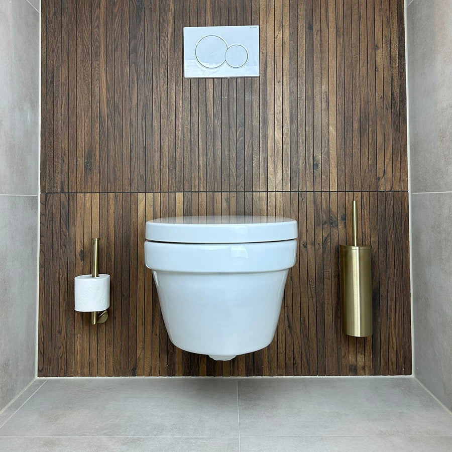 Een moderne badkamer toont een aan de muur gemonteerd wit toilet tegen houten lambrisering, met rechts de Fabiano Gold WC-set van yessential.nl, inclusief een gouden toiletborstelhouder. De vloer en zijwanden zijn lichtgrijs betegeld.