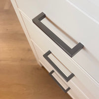 Close-up van drie witte lades met moderne Vasco Gunmetal handgrepen van yessential.nl, tegen een lichte houten vloer. De lades hebben een lichte hoek, waardoor de strakke lijnen en het minimalistische ontwerp goed tot hun recht komen.