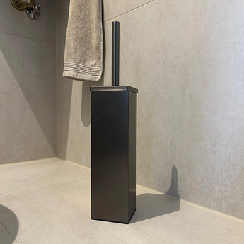 De Cano Gunmetal WC Set van yessential.nl heeft een moderne rechthoekige zwarte houder met een gunmetal handvat, ideaal voor je toiletaccessoireset. Hij is no-drill en past netjes op lichte tegels bij een beige handdoek.