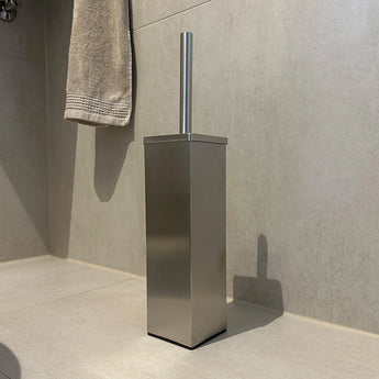 De Velasco RVS toiletborstel met houder van yessential.nl, met rechthoekig design, staat op een lichtgekleurde tegelvloer in de badkamer nabij een beige handdoek aan de muur.