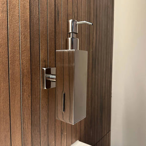 Het Sonego Chrome zeeppompje van yessential.nl is een moderne, muurbevestigde RVS dispenser met pompdeksel en een venster om het zeepniveau te tonen, gemonteerd aan een houten badkamerpaneel.