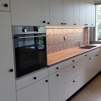 Een moderne keuken met witte kasten met de Adaro Black Knob van yessential.nl, een ingebouwde oven, een houten aanrechtblad, verlichting onder de kasten, een achterwand met patroontegels, een roestvrijstalen gootsteen en een groot raam.