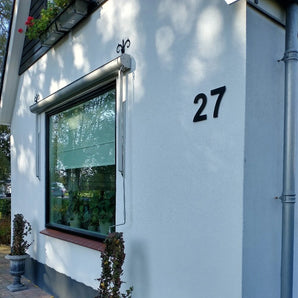 Een wit huis met een groot raam, zwarte luiken en yessential.nl Animo Black 15cm (geen boor) plakletters in vet nummer 27 op de buitenmuur. Een kleine potplant staat onder het raam bij de hoek.