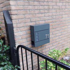 De Nevio Anthracite brievenbus van yessential.nl met nummer 71 is gemonteerd op een bakstenen muur, naast groene planten en een zwart metalen hek.