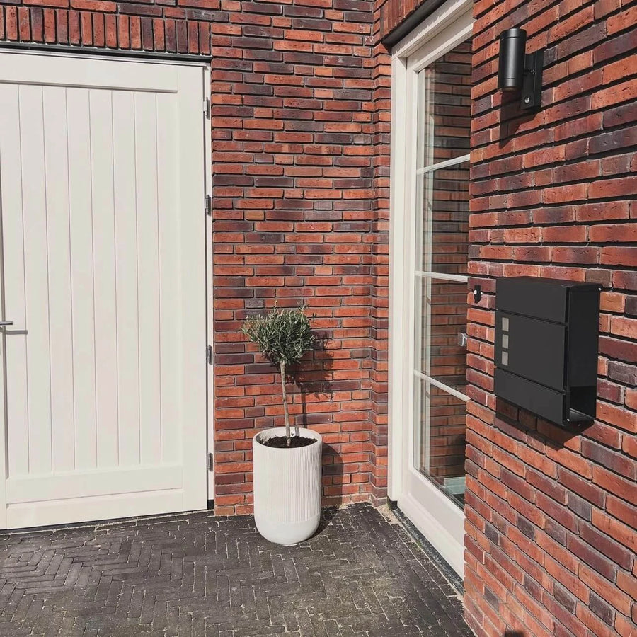 Een modern bakstenen huis met een witte deur, groot raam, Nevio Antraciet brievenbus van yessential.nl, een kleine boom in pot in een witte plantenbak en een zwarte buitenlamp.
