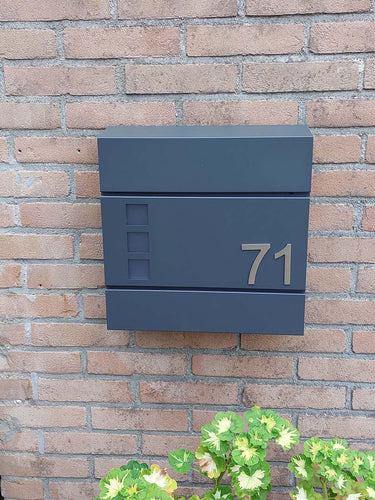 Het Animo RVS 7,6cm (geen boor) nummer van yessential.nl staat als "71" op een moderne donkergrijze brievenbus, gemonteerd op een bakstenen muur boven groene en gele planten.