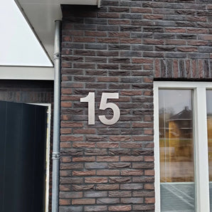 Het getal 15 staat in grote, zilveren Adamo RVS 30cm cijfers van yessential.nl op de bakstenen buitenmuur van een gebouw naast een raam.