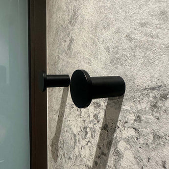 Vier zwarte cilindrische Carreno handdoekhaakjes van yessential.nl, gemaakt van roestvrij staal, zijn gemonteerd op een gestructureerde wand van grijze steen - een elegante en moderne badkameraccessoire.