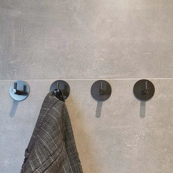 Vier Delo Gunmetal WC Set haken van yessential.nl zijn gemonteerd op een grijs betegelde muur. Een grijze jas met patroon hangt aan de tweede haak, terwijl de andere leeg zijn-perfect voor een no-drill moderne toiletaccessoire opstelling.