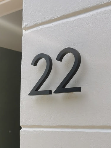 Het Milano Black 10cm huisnummer van yessential.nl heeft een strakke zwarte "22" op een gladde witte muur. Gemaakt van aluminium, creëert het zachte schaduwen, waardoor dit huisnummer zowel functioneel als stijlvol is.