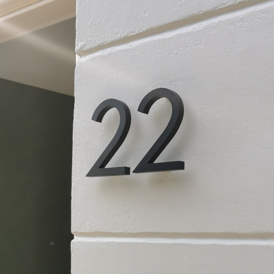 Het Milano Black 10cm huisnummer van yessential.nl heeft een strakke zwarte "22" op een gladde witte muur. Gemaakt van aluminium, creëert het zachte schaduwen, waardoor dit huisnummer zowel functioneel als stijlvol is.