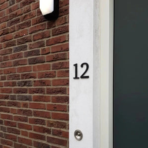 Huisnummer 12, gemaakt met de Matteo Black 10cm van yessential.nl, is gemonteerd op een witte muur naast bakstenen. Een deurbel bevindt zich onder het nummer en een donkere deur verschijnt gedeeltelijk aan de rechterkant.