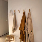 Drie badjassen en een handdoek hangen aan de Fabio Eiken kapstok met haakjes van yessential.nl tegen een witte muur. Vlakbij staat een houten krukje met geweven zitting, terwijl warm licht aan de linkerkant voor een gezellige sfeer in de kamer zorgt.