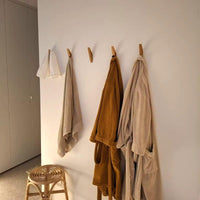 Drie badjassen en een handdoek hangen aan de Fabio Eiken kapstok met haakjes van yessential.nl tegen een witte muur. Vlakbij staat een houten krukje met geweven zitting, terwijl warm licht aan de linkerkant voor een gezellige sfeer in de kamer zorgt.