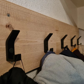 Een rij van 10x Huso Black haken van yessential.nl is gemonteerd op een lichte houten plank en houdt jassen en jacks tegen een witte muur met textuur voor een strakke, moderne look.