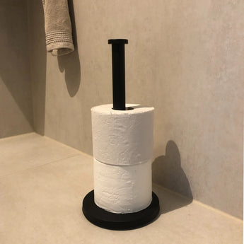 Een Jurado Black toiletrolhouder van yessential.nl staat verticaal in de hoek van een beige betegelde badkamer, met daarin twee witte toiletrollen, met daarboven een netjes opgevouwen handdoek aan de muur.