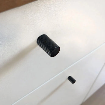 Close-up van twee Remo Black Deluxe knoppen van yessential.nl op een witte kast, waarbij hun getextureerde ontwerp subtiele schaduwen werpt. De onscherpe achtergrond vestigt de aandacht op deze moderne kastknopdetails.