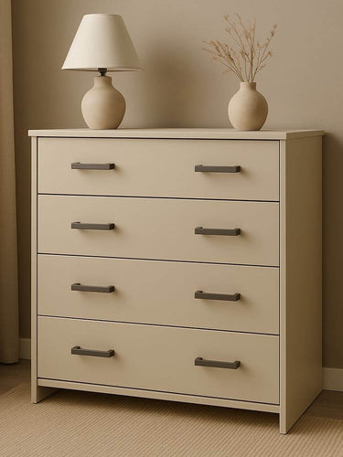 De Vasco Gunmetal commode van yessential.nl heeft vier lades met gunmetal handgrepen en staat in een neutrale kamer. Bovenop staan een crèmekleurige lamp en een vaas met gedroogde takken, geplaatst bij een raam met lichte gordijnen.