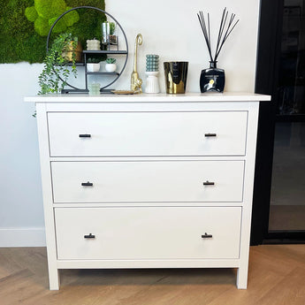 Een witte ladekast met drie lades en Remo Black knoppen van yessential.nl staat op een houten vloer. Vazen, planten, kaarsen en een ronde spiegel staan erop en op de wandplank erboven.