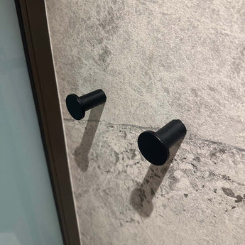 Vier Carreno Black handdoekhaakjes van yessential.nl zijn gemonteerd op een getextureerde lichtgrijze stenen muur met schaduwen door schuin licht. Links is een verticaal metalen frame zichtbaar-ideale badkamer accessoires.
