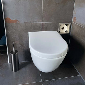 Een modern wit wandcloset met een gesloten deksel, naast de Tygo Gunmetal WC Set (no-drill) van yessential.nl en een vierkante verzonken toiletpapierhouder in een badkamer met donkergrijze betegelde muren en vloer.