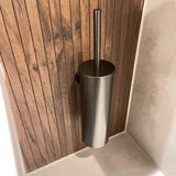 De Lauro Gunmetal WC-set van yessential.nl is een slanke metalen toiletborstelhouder met een verticale handgreep, gemonteerd op een wand met houten panelen naast lichtgekleurde tegels, perfect voor luxe badkamer accessoires collecties.